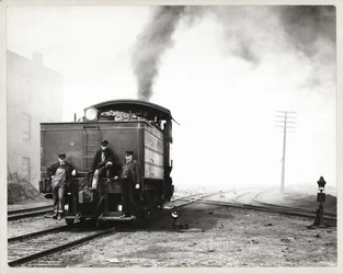 Drei Männer auf dem Rücken einer Rangierlokomotive in einem unbekannten Rangierbahnhof